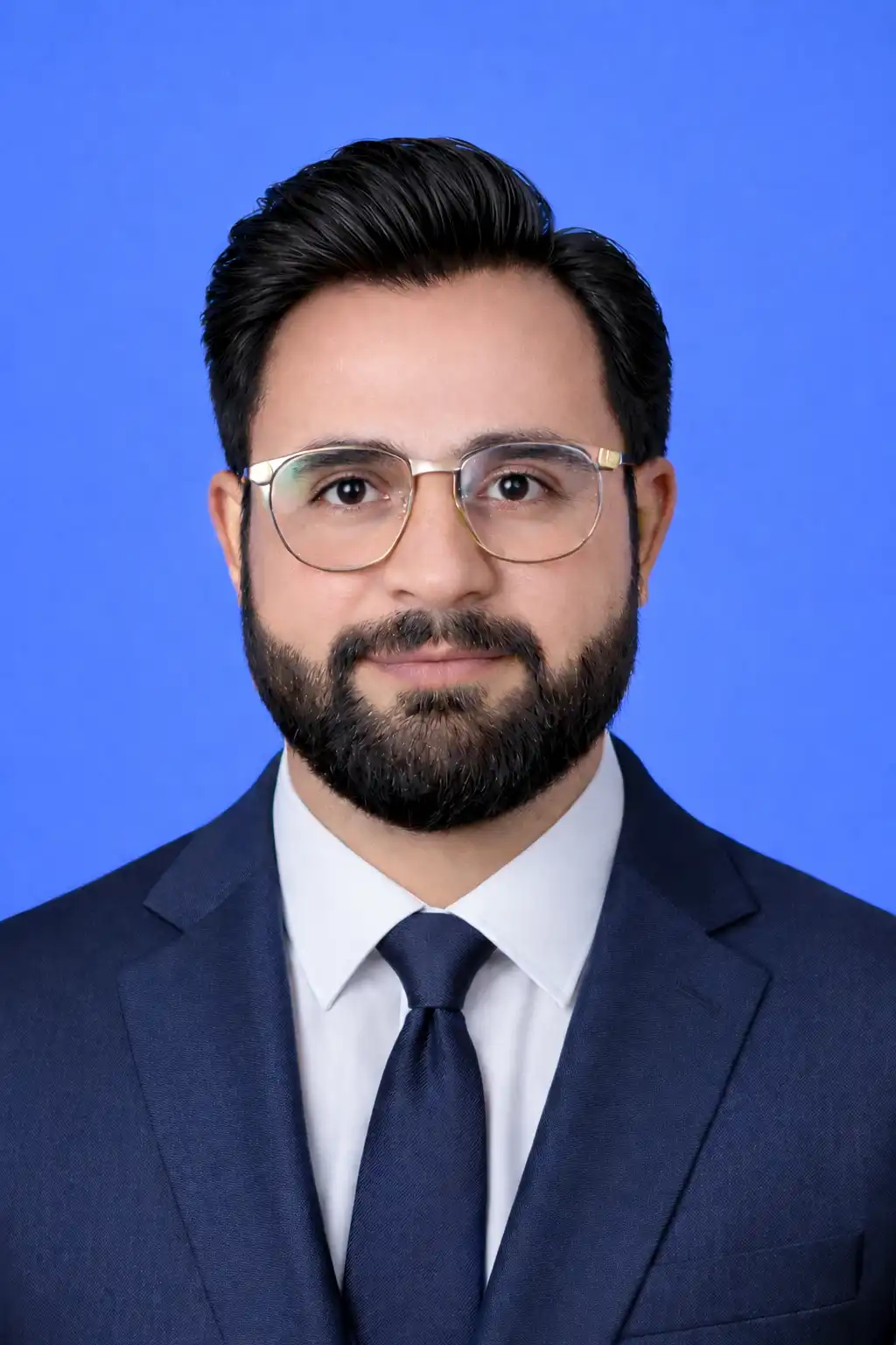 Professional headshot: man with dark hair and beard wearing glasses, a navy suit and blue tie against a blue background.