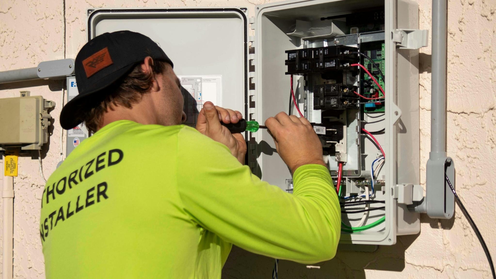 Electrical technician in a neon shirt tightening wires inside an outdoor electrical panel mounted on a stucco wall