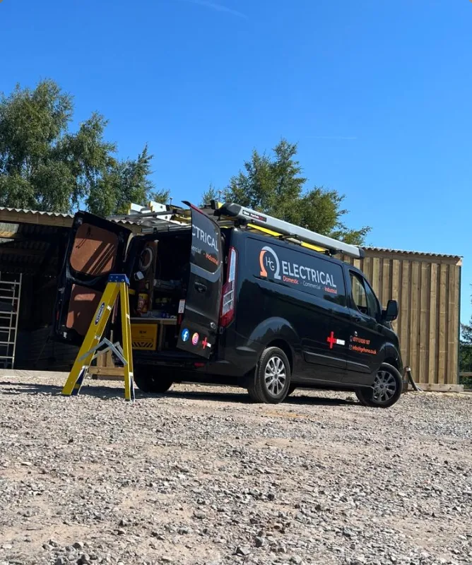 Black electrical service van on a gravel lot, ladder on the roof and tools visible in the open rear doors near a wooden shed.