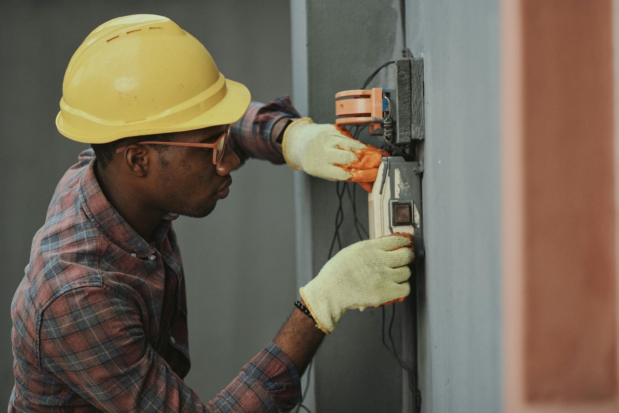 Construction worker wearing a yellow hard hat and gloves, wiring an electrical panel on a wall.